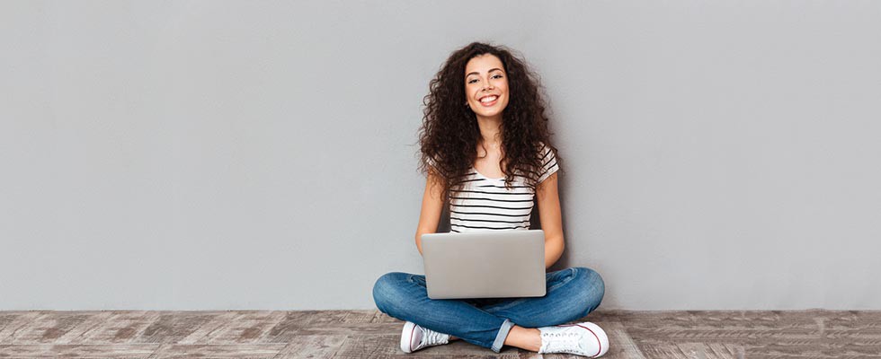 Portrait of satisfied female with beautiful smile enjoying watching movie in silver computer and sitting in lotus pose on the floor over grey wall