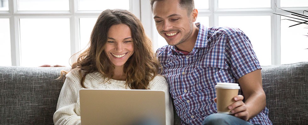Smiling young couple enjoying morning coffee with computer, sitting on sofa at home together; happy man and woman laughing looking on laptop screen having fun while programming
